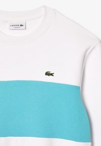 Sudadera blanca con una franja horizontal turquesa. Presenta un cuello redondo de canalé y un pequeño logo de cocodrilo bordado en verde. Diseño de ajuste clásico.