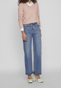 Ljusrosa v-ringad tröja, i kombination med högmidjade blå vida jeans. Modellen bär krämfärgade plattformsskor. Enhetlig bakgrund.