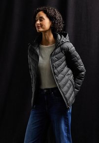 Svart pufferjacka med huva, med chevron-stygn, en dragkedja framtill och sidofickor, bärs över en ljus tröja och blå jeans.