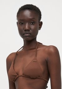 Smeđa trokutasta bikini s teksturiranim materijalom, tankim naramenicama i logotipom. Jednostavan dizajn naglašava minimalno pokrivanje i bešavna izrada.