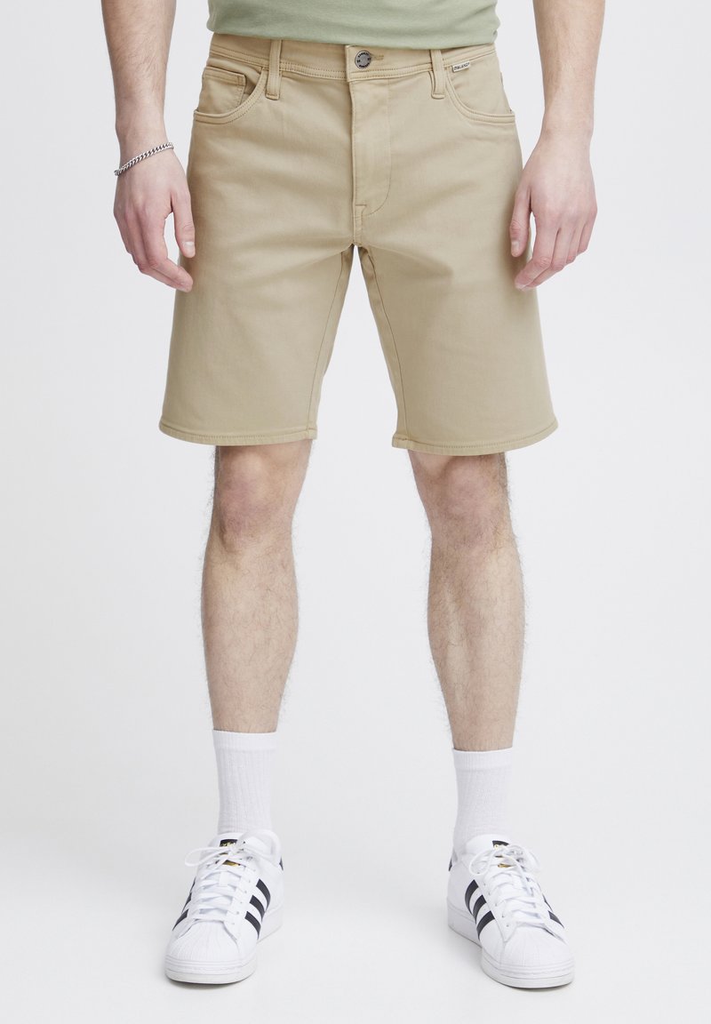 Pantalones cortos de algodón beige con cierre de botón frontal, cinco bolsillos y detalles de costura. Llevados con calcetines blancos y zapatillas de deporte de rayas negras.