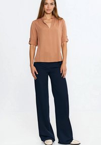 Kobieta ubrana w brązową bluzkę z dekoltem w serek i podwiniętymi rękawami, granatowe spodnie o szerokich nogawkach oraz beżowe mokasyny, stojąca na tle jednolitego białego tła.