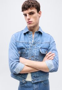 Νέος άνδρας με σταυρωμένα χέρια, φορώντας ανοιχτό μπλε τζιν μπουφάν και τζιν παντελόνι, μπροστά σε απλό ανοιχτό φόντο.