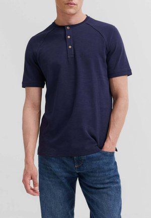 Uomo che indossa una maglietta henley blu navy a manica corta con tre bottoni di legno e jeans di denim blu, in piedi davanti a uno sfondo uniforme.