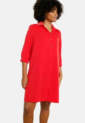 Femme portant une robe chemise rouge vif, arrivant aux genoux, avec des manches trois-quarts et un col, debout devant un fond blanc uni.