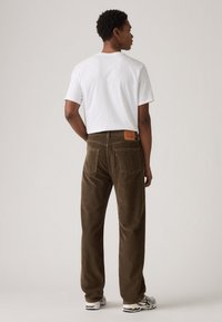 Hombre en pantalones de pana marrón, zapatillas blancas y una camiseta blanca simple, de pie de espaldas frente a un fondo liso.