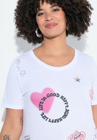 T-shirt blanc à manches courtes avec un cœur rose et un texte noir "LA VIE EST BELLE HEUREUX" accompagné d'étoiles colorées et d'accents graphiques.