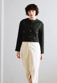 Giacca corta nera con texture, chiusura a bottoni e maniche lunghe, abbinata a una gonna avvolgente beige con spacchi frontali.