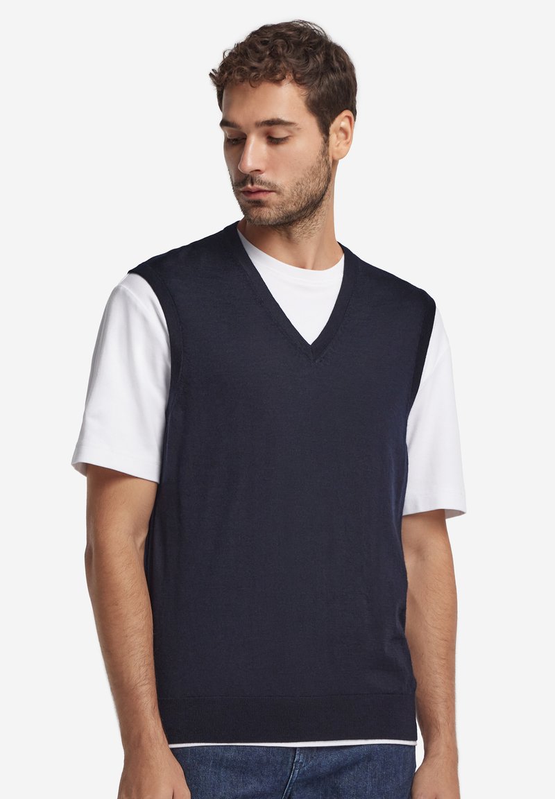 Marinblå stickad väst med V-ringning, bärs över en vit t-shirt. Texturen är mjuk med ribbade kanter och en avslappnad passform.