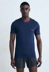 T-shirt azul-marinho de manga curta, feito de um tecido suave, apresentando um logo vermelho "HUGO" no lado esquerdo do peito. Design simples e ajustado.