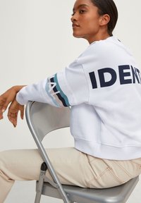 Persoon die zijwaarts op een metalen stoel zit, met een witte sweatshirt aan waarop de tekst "IDENT" op de rug staat en een gestreept ontwerp op de mouwen.