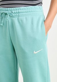 Lys tealkfargede joggebukser laget av myk stoff med elastisk midjebånd og snøring, med en liten hvit Nike-logo på siden.