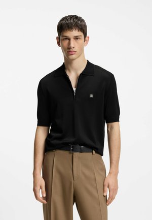 Junger Mann trägt ein schwarzes kurzärmeliges Polohemd mit Reißverschluss und kleinem Logo, sandfarbene Hose und schwarzen Gürtel vor weißem Hintergrund.