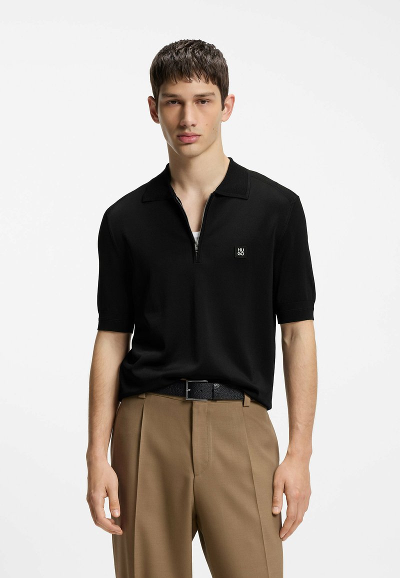 Jeune homme portant un polo noir à manches courtes avec fermeture éclair et petit logo, pantalon beige et ceinture noire sur fond blanc uni.