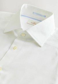Hvid bomuldsbluse med en struktureret overflade, spids krave og gule knapper. Mærket viser "NEXT SIGNATURE" med en lyseblå accent.