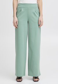 Lichtgroene wijde broek van zachte stof, met een hoge tailleband en voorplooien, gecombineerd met dunne zwarte sandaaltjes met bandjes.