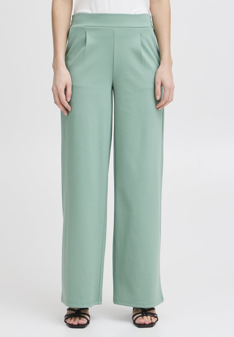 Lichtgroene wijde broek van zachte stof, met een hoge tailleband en voorplooien, gecombineerd met dunne zwarte sandaaltjes met bandjes.