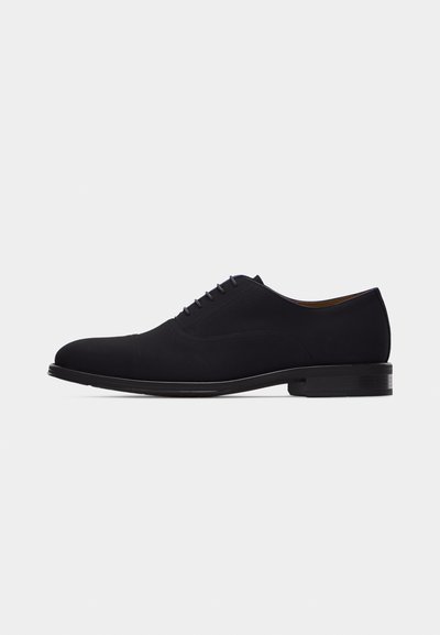Chaussure habillée Oxford en cuir noir avec laçage fermé, bout droit et talon bas empilé sur fond blanc.