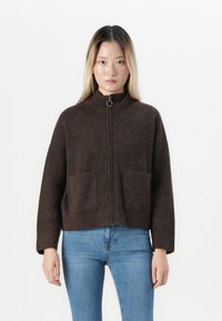 Femme portant un sweat à zip marron foncé avec des poches et un jean bleu clair, se tenant contre un fond blanc uni.