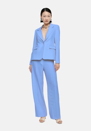 Donna in piedi che indossa un completo su misura azzurro chiaro con pantaloni a gamba larga, blazer aperto, collana d'argento, orecchini vistosi e tacchi beige con punta aperta.