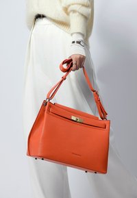 Orange läderhandväska med en strukturerad design, utrustad med ett guldfärgat spänne, justerbar rem och subtil märkning på framsidan.