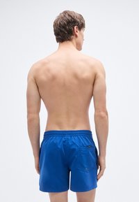 Μπλε μαγιό με ελαστική μέση, πίσω τσέπη και λεία υφή, εμφανιζόμενο από πίσω,  σε απλό φόντο.