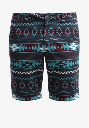 Mann trägt ein hellblaues Kurzarm-Denimhemd, gemusterte knielange Shorts in Türkis, Schwarz, Rot und rote Sneakers mit weißen Schnürsenkeln.