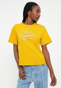 Kollane puuvillane t-särk lühikeste varrukatega, millel on retrostiilis trükitud "New Balance" logo kontrastse värviga ülemise rinnaosas.