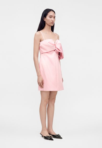 Femme aux longs cheveux noirs portant une robe rose sans manches avec un grand nœud et des chaussures plates noires pointues, debout de côté.