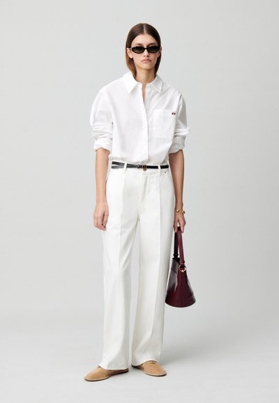 Camicia bianca con bottoni e tasca sul petto, pantaloni bianchi a vita alta, scarpe beige e borsa rossa scura. Il modello indossa occhiali da sole neri.
