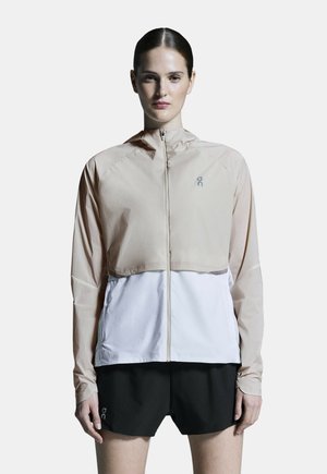 Giacca a vento leggera con la parte superiore beige e la parte inferiore bianca, dotata di zip frontale e dettagli del logo discreti. Abbinata a pantaloni corti neri.