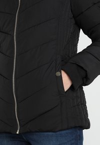 Svart puffjacka med ett chevron-quiltat mönster, silverdragkedja och ribbade mudd. Har sidofickor och en stilren design.