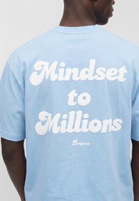 Osoba nosi svetlomodré tričko s tučným bielym textom „Mindset to Millions“ a menším logom „Dropsize“ na chrbte.