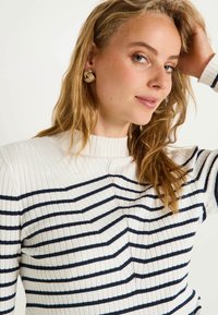 Pull côtelé à rayures avec un col montant en crème et bleu marine, présentant un design texturé et des motifs en zigzag subtils. Accent avec des boucles d'oreilles dorées.