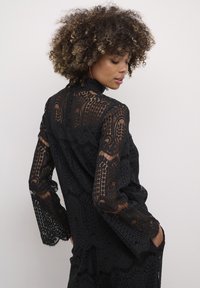 Black lace top featuring intricate patterns, long sleeves, and a high neckline. Fabric is sheer with textured details and scalloped edges.