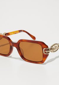 Ochelari de soare cu tentă maro, cu un cadru gros din scoică țestoasă și accesorii din metal auriu, având un accent decorativ sub formă de legătură pe tâmple.