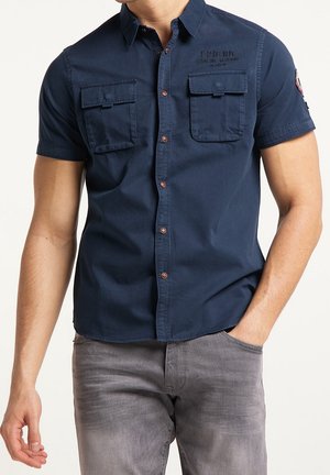 Mężczyzna ubrany w granatową koszulę z krótkim rękawem zapinaną na guziki, z dwoma kieszeniami na piersi zapinanymi na guziki oraz szare dżinsy, jedną ręką w kieszeni, na jednolitym jasnym tle.