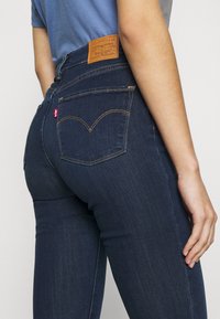 Jeans de tiro alto de color azul oscuro, fabricados en denim, con un parche de marca de cuero, costuras en contraste y dos bolsillos traseros con un diseño curvado.