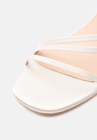 Gros plan sur une sandale en satin blanc avec de fines lanières croisées et un bout carré sur un fond clair uni.