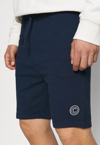 Laivastonsiniset puuvillahousut, joissa on joustava vyötärönauha, säädettävä kiristysnauha ja vasemmassa lahkeessa pyöreä logoplässi. Tasainen pinta, polvenpituinen.