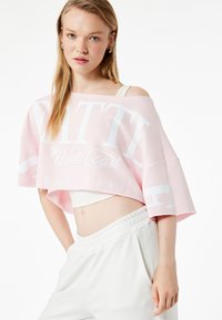 Sudadera rosa corta con texto impreso en blanco "SEATTLE West Coast EST. 1991" y un ajuste relajado. Llevada sobre una camiseta sin mangas blanca y pantalones con cintura elástica.