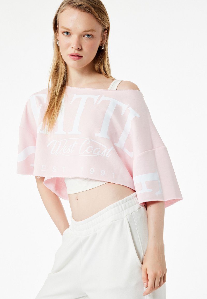 Sudadera rosa corta con texto impreso en blanco "SEATTLE West Coast EST. 1991" y un ajuste relajado. Llevada sobre una camiseta sin mangas blanca y pantalones con cintura elástica.