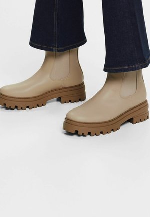 Beige ankelstøvler med chunky brune såler, båret med mørkeblå viddebukser mod en ensartet hvid baggrund.