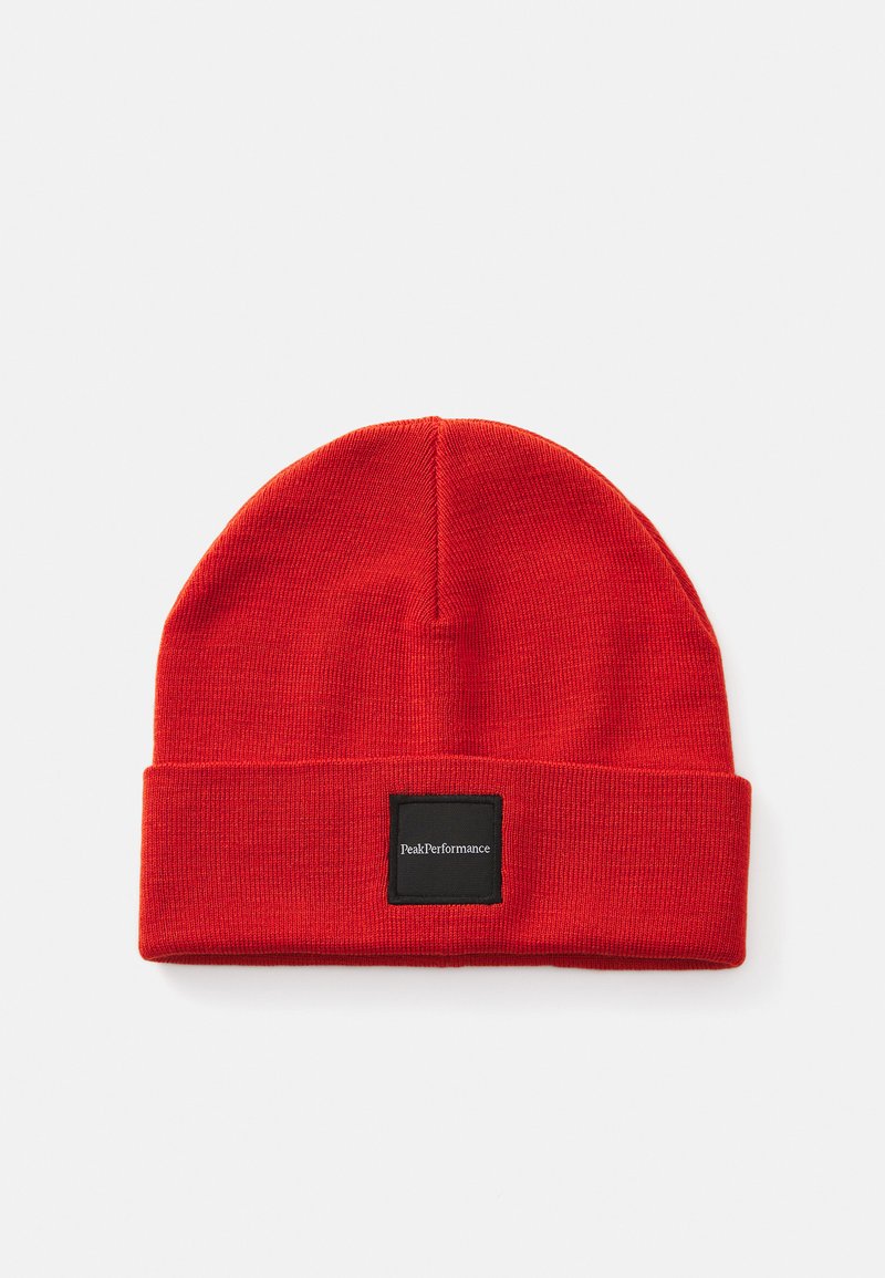 Red knit beanie with a folded cuff. Features a square black label with white text. Soft texture; rounded top with smooth seams.