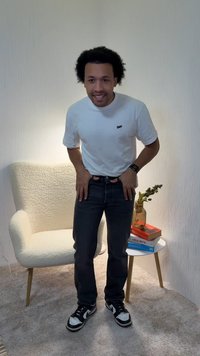 White short-sleeve shirt, dark jeans, and black sneakers with white accents. Beige textured chair and a small table with books and a vase.