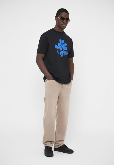 T-shirt en coton noir avec motif graphique bleu, associé à un pantalon en denim beige et des baskets noires. Modèle portant des lunettes de soleil.