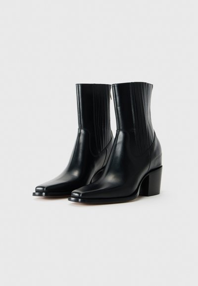 Ett par eleganta svarta läderankelboots med klackar och vertikala sömnadsdetaljer på sidorna, presenterade mot en enkel bakgrund.