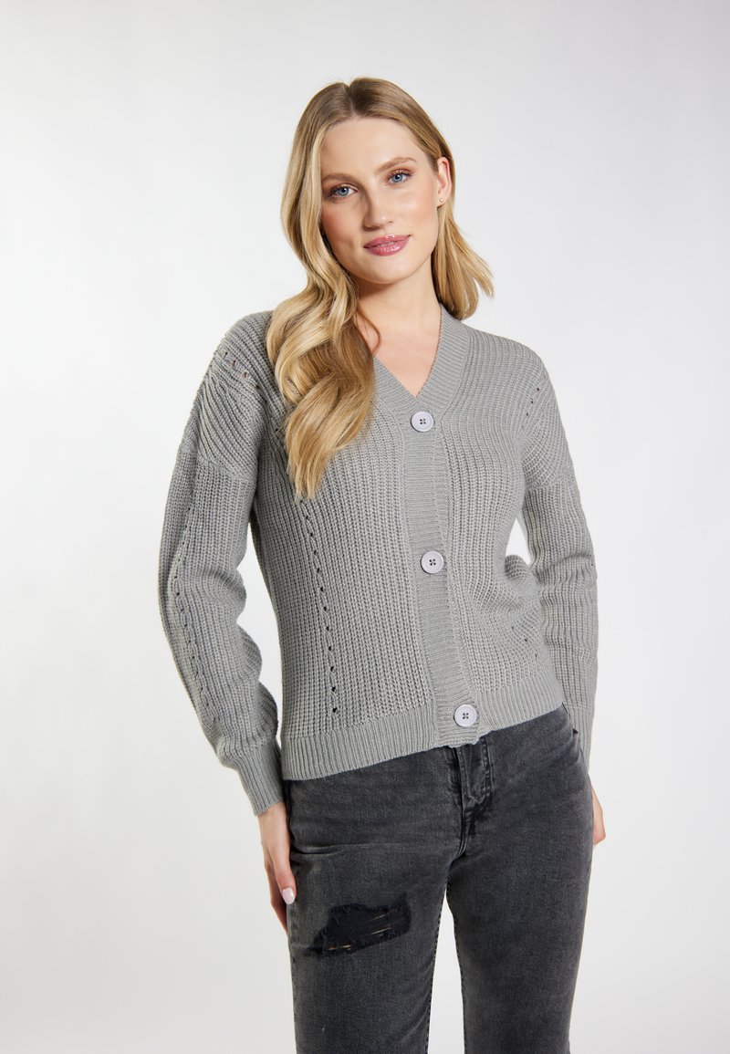 Cárdigan gris de punto con escote en V, mangas largas, tres botones grandes y detalles de calados a lo largo de las mangas. Combinado con jeans negros desgastados.