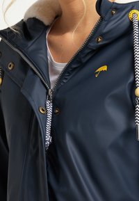 Marinblå vattentät jacka med en mjuk krämfärgad krage, silverdragkedja och gul logotyp. Har mönstrade dragsnören och metallaccenter.