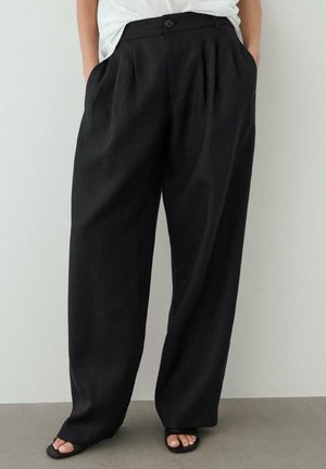 Osoba w luźnych czarnych spodniach z wysokim stanem z plisami, ręce w kieszeniach, w połączeniu z czarnymi sandałami na obcasie z odkrytymi palcami i białą bluzką.